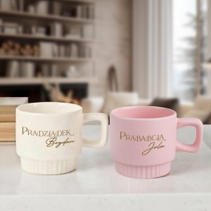 Dwie filiżanki stoją na blacie, jedna biała z napisem 'Pradziadek Bogdan', a druga różowa z napisem 'Prababcia Jola', w tle szyby i regał z książkami oraz drewnianymi elementami dekoracyjnymi.
