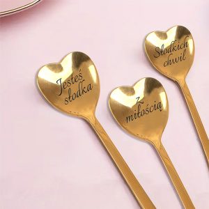 lyzeczki-na-walentynki-serca-z-grawerem.jpg Trzy złote łyżeczki w kształcie serca z napisem: 'Jesteś słodka', 'Z miłością', i 'Słodkich chwil'. Tło różowe.