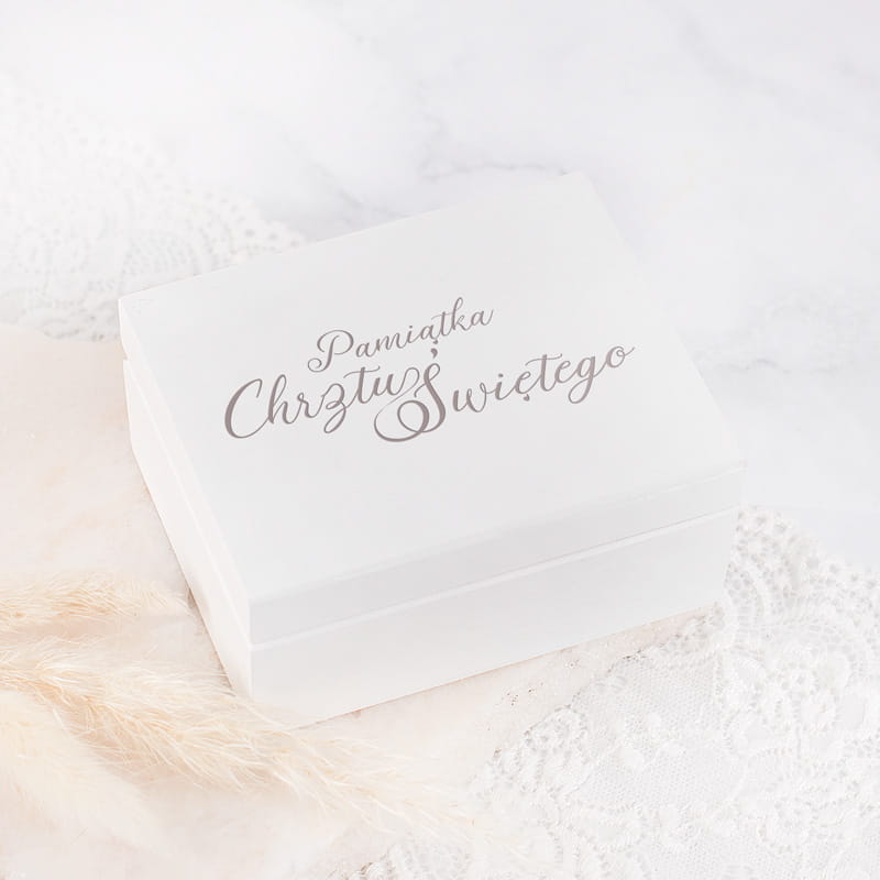 Biała, elegancka skrzynka z napisem "Pamiątka Chrztu Świętego" w stylu kaligraficznym, umieszczona na jasnym tle ozdobionym koronką i suszonymi kwiatami.
