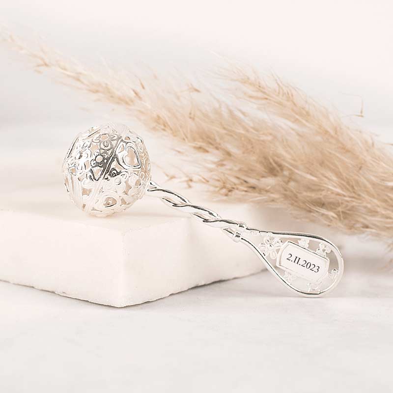 Elegancka, srebrna łyżeczka z ozdobną, ażurową kulą na końcu, umieszczona na jasnym tle, obok delikatnej trawy pampasowej; na łyżeczce widoczna jest data: 2.11.2023.