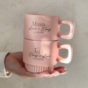 Dwie różowe filiżanki ułożone jedna na drugiej, z napisem 'MAMA' na górnej i 'TATA' na dolnej, z imionami 'Liwi i Eleny' oraz datą '02.01.2026'. Filiżanki trzymane w jednej ręce na tle jasnej ściany.