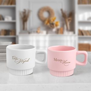 personalizowany-kubek-z-ryflowaniem-dla-rodzicow.jpg Dwie filiżanki na blacie: jedna biała z napisem 'Tata Wojtek' w złotym kolorze, druga różowa z napisem 'Mama Maria' także w złotym kolorze, w tle jasne wnętrze z regałem pełnym książek.