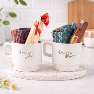 Dwa białe kubki na blacie, jeden z napisem 'Babcia Jola' i drugi z 'Dziadek Bogdan', wypełnione kolorowymi skarpetami i słodyczami, na okrągłej podstawce z jasnego materiału, otoczone delikatnymi kwiatami.