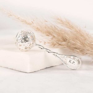 Srebrna łyżeczka z misternie zdobioną kulką na końcu, leżąca na jasnym, prostokątnym kawałku materiału, z logo 'K.P.' na trzonku, w tle delikatna trawa pampasowa.