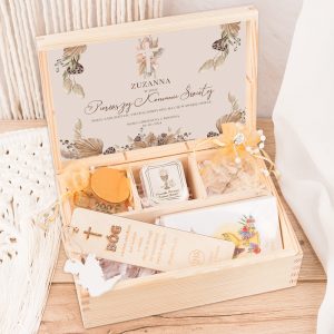 W drewnianym pudełku z delikatnym nadrukiem znajduje się zestaw upominkowy z okazji Pierwszej Komunii Świętej dla Zuzanny. W środku znajdują się różne przedmioty, w tym świeca, którego etykieta jest ozdobiona złotą grafiką, wosk, kartka oraz zakładka do książki z haftem w kształcie krzyża.