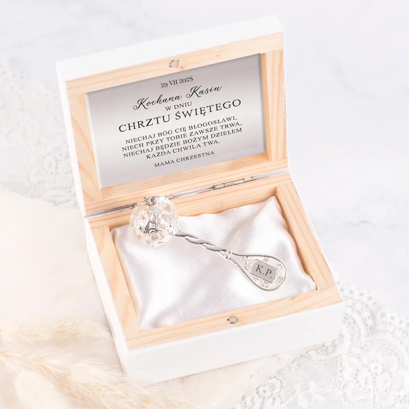 Eleganckie pudełko zawierające srebrną łyżeczkę z dekoracyjną kulą oraz tekstem dedykacyjnym na wieczku, witające dziecko z okazji chrztu. Stitch, tkanina w środku pudełka i ozdobne detale widoczne w tle.