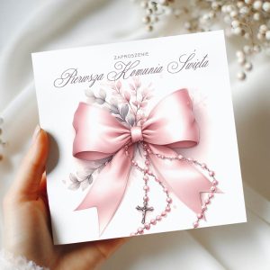 Eleganckie zaproszenie na Pierwszą Komunię Świętą, ozdobione różową wstążką i różowymi koralikami z krzyżykiem, trzymane w dłoni na tle jasnej tkaniny.