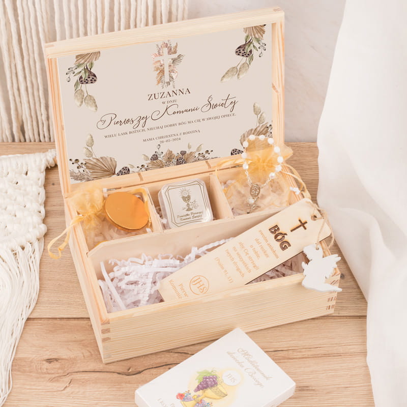 Drewniana skrzyneczka z prezentem na Pierwszą Komunię Świętą, zawierająca kartkę z imieniem Zuzanna, słodkości, medalik oraz zakładkę z modlitwą. Ozdobiona delikatnymi kwiatowymi motywami i krzyżem.