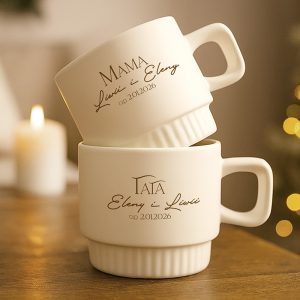 Dwie biały kubki ceramiczne ustawione jeden na drugim. Górny kubek ma napis 'MAMA' oraz daty, a dolny 'TATA' z datami. Tło jest rozmyte z delikatnym światłem świecy i ozdobnymi światełkami.