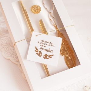 Zestaw złotych akcesoriów do Pierwszej Komunii Świętej w eleganckim pudełku, zawierający dwie pióra i okrągłą pieczęć, z kartką z napisem 'Pierwsza Komunia Św. Franka'.