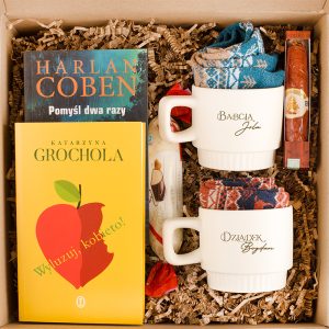 Zestaw prezentowy w pudełku, zawierający dwie filiżanki z imionami 'Babcia Jola' i 'Dziadek Bogdan', dwie książki, w tym 'Pomysł dwa razy' Harlana Cobena i 'Wyluzuj, Kobieto!' Katarzyny Grocholi, a także słodycze i ciepłe skarpetki.