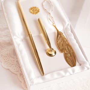 zlota-zakladka-i-dlugopis-na-komunie-dla-dziecka Zestaw złotych przyborów do pisania, w tym długopis, stalówka oraz ozdobny przycisk do papieru w kształcie pióra, umieszczony na białym tle z miękkim materiałem.