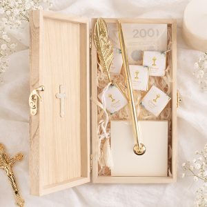 Drewniane pudełko z zawartością: złoty długopis, kartki oraz małe opakowania z cukierkami, otoczone dekoracyjnymi wstążkami.