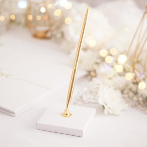 Złoty długopis umieszczony na białym podstawku, otoczony dekoracjami i jarzącymi się światełkami.