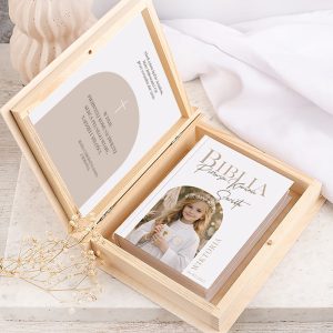 Otwarte drewniane pudełko zawierające Biblię z okładką przedstawiającą dziewczynkę w białej sukience i wianku, z instruckją wewnętrzną.