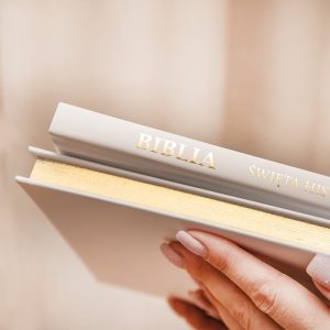 Osoba trzymająca książkę z widocznym tytułem 'Biblia' oraz podtytułem 'Święta Historia' na okładce, w tle jasne, neutralne tło.
