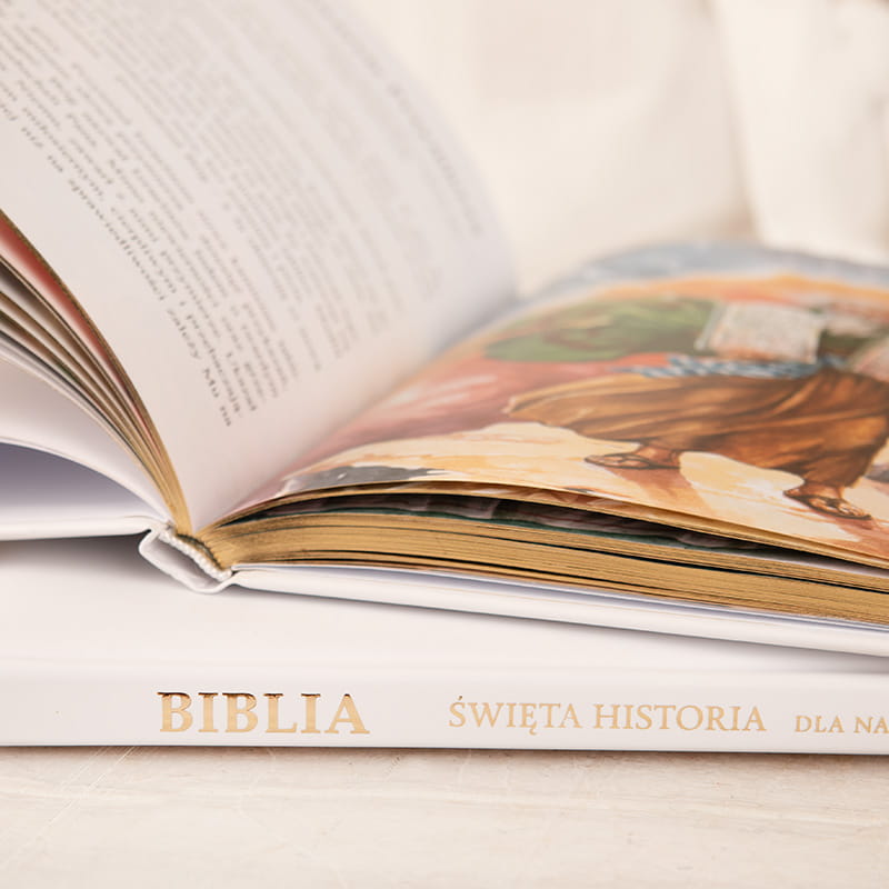 Otwarta Biblia z ilustrowaną stroną, na której widoczna jest postać w szatach, trzymająca kartę, z widocznymi złotymi brzegami stron; tytuł na okładce to "BIBLIA ŚWIĘTA HISTORIA DLA NAS".