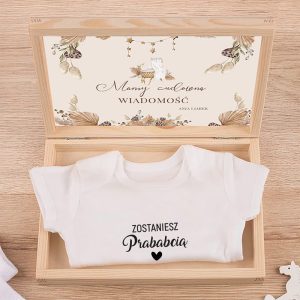 Biały bodziak z napisem "Zostaniesz Prababcią" leży w drewnianym pudełku z ozdobnym wnętrzem.