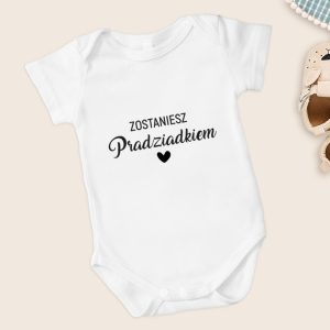 Biały body dziecięcy z napisem "Zostaniesz Pradziadkiem" oraz serduszkiem. Obok widoczne buciki dziecięce.