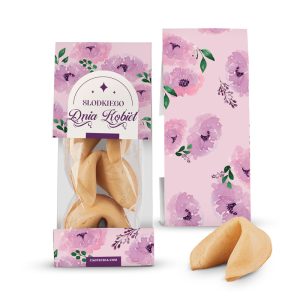 Opakowanie ciastka z wróżbą z różowym motywem kwiatowym, w środku trzy ciastka widoczne przez przezroczystą szybkę, obok jedno ciastko na blacie.