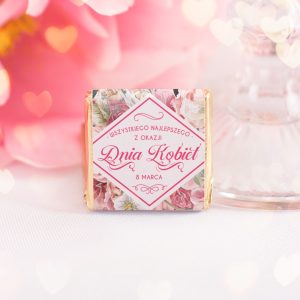 Mała czekoladka w ozdobnym opakowaniu z różowymi kwiatami, napisem 'Wszystkiego najlepszego z okazji Dnia Kobiet 8 Marca', na tle rozmytego różowego tła z serduszkami.