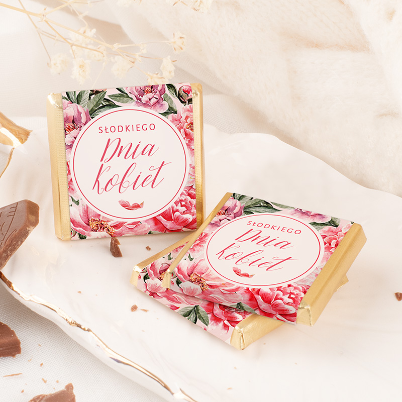 Dwa kawałki czekolady na talerzu z kremowym tłem, z opakowaniem ozdobionym kwiatami i napisem "Słodkiego Dnia Kobiet" na górze, położone obok kawałka czekolady.