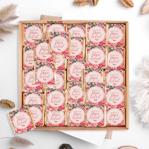 Elegancka pudełko z różnorodnymi małymi czekoladkami, każda z ozdobną etykietą w stylu rustykalnym z różami, w tle kilka dekoracyjnych elementów, takich jak suszone rośliny i orzechy.