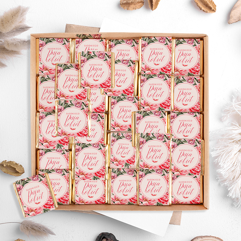 Elegancka pudełko z różnorodnymi małymi czekoladkami, każda z ozdobną etykietą w stylu rustykalnym z różami, w tle kilka dekoracyjnych elementów, takich jak suszone rośliny i orzechy.