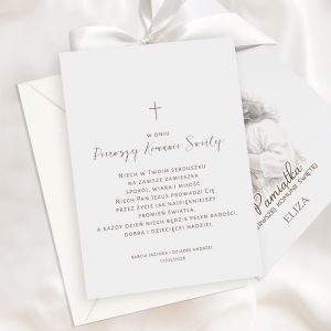 Elegancka karta zaproszeniowa na Pierwszą Komunię Świętą z delikatnym ornamentem krzyża i złotym tekstem, z życzeniami spokoju, wiary i miłości, przypinana w białą kopertę z kokardą.