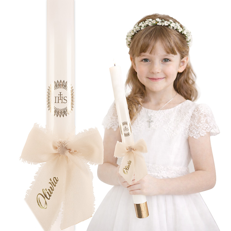 Dziewczynka w białej sukience z koroną na głowie trzyma świecę z ozdobną wstążką, na której widnieje imię „Olivia”.