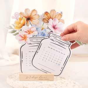 kalendarz-bukiet-dla-mamy Stojący kalendarz z rysunkiem słoika i kolorowymi kwiatami, przytrzymywany ręką, z miesiącem marzec 2026.