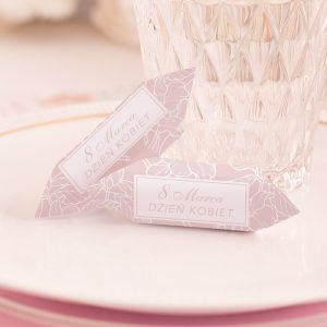 Dwa ozdobne bilety na stół z napisem "8 Marca Dzień Kobiet" w różowym kolorze, leżące obok przezroczystego szklanki na eleganckim talerzu.