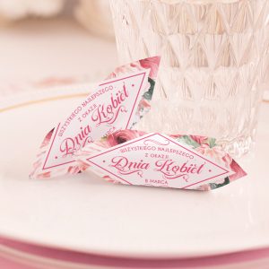 Dwie ozdobne kartki z napisem "Dnia Kobiet" oraz datą "8 Marca", leżące na białym talerzu obok szklanki z wodą. Kartki ozdobione różami.