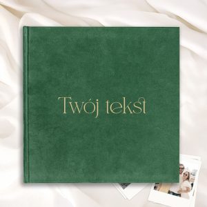 Zielona okładka książki z napisem "Twój tekst" w złotym kolorze, leżąca na jasnym tle z fałdami materiału i zdjęciami.