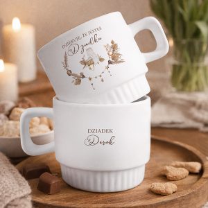 Dwie białe filiżanki, jedna ustawiona na drugiej, z dekoracyjnymi napisami i wzorami na boku. W tle świece i wypieki.