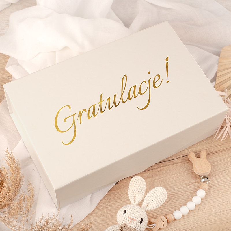 Biały pudełko z napisem "Gratulacje!" w złotym kolorze, otoczone jasnymi materiałami i dekoracjami.