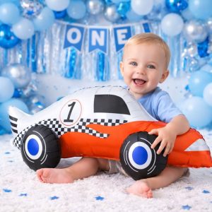 balon-na-pierwsze-urodziny.jpg Małe dziecko w niebieskiej koszulce siedzi na dywanie i uśmiecha się, trzymając w rękach nadmuchiwany samochód wyścigowy. W tle dekoracje urodzinowe.