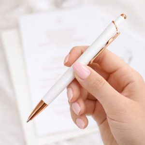 Biała długopis z różowym złotem trzymany w ręce, w tle lekko rozmyte strony dokumentów.