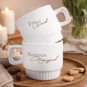 Dwie białe filiżanki z złotym imieniem i "Najlepszy Nauczyciel", ustawione jedna na drugiej na drewnianym stole.