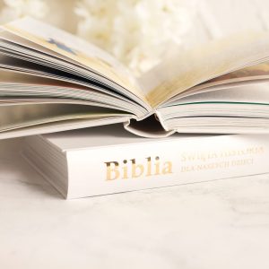 biblia-na-komunie-dla-chlopca.jpg Otwarta Biblia z ilustracjami skierowana do dzieci, leżąca na białym blacie, z delikatnymi kwiatami w tle.