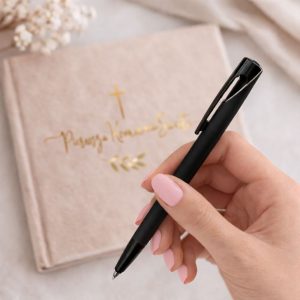 czarny-dlugopis-elegancki.jpg Czarna długopis trzymany w ręce nad eleganckim, jasnym zeszytem z złotym napisem.