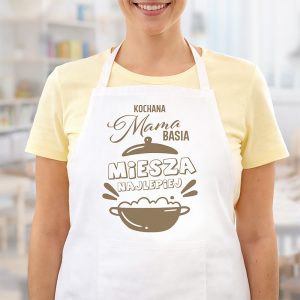 Fartuch kuchenny z napisem "Kochana Mama" oraz "Miesza Najlepiej", w kolorze białym, noszony przez osobę w żółtej koszuli.
