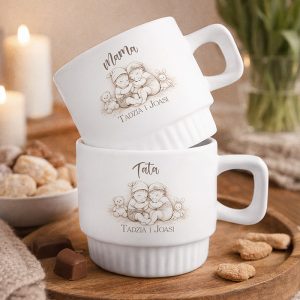 Dwie białe filiżanki z ilustracjami dzieci i napisami "Mama" oraz "Tata", ustawione na drewnianym blacie obok słodkości.