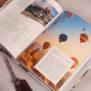 ksiazka-z-bucket-list-na-komunie.jpg Otwarte czasopismo z fotografią kolarza górskiego oraz balonami na gorące powietrze unoszącymi się nad górami.