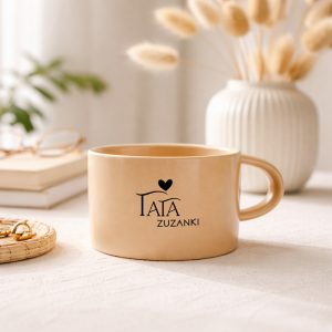 Beżowy kubek z logo "TATA" w otoczeniu dekoracyjnych roślin i akcesoriów na stole.