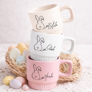 Trzy kubki w pastelowych kolorach z rysunkami króliczka i imionami: Natalia, Oliwia i Amelia, ustawione w koszyku z kolorowymi jajkami.