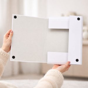 Osoba trzyma otwartą białą teczkę w miękkim pomieszczeniu, z widoczną pustą stroną oraz kieszonkami na dokumenty.