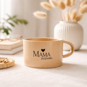 Jasny kubek z napisem "MAMA", stojący na stole obok biżuterii i dekoracji.
