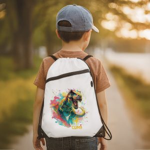 plecak-dla-dziecka.jpg Chłopiec z plecakiem w kolorze białym, z ilustracją dinozaura, stoi na drodze w parkowym otoczeniu.