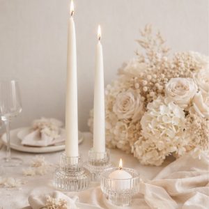 Dwa białe świece w szklanych świecznikach obok małej świecy, wszystko otoczone jasnymi kwiatami i delikatnym obrusem.